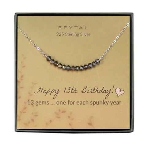 grey jewelry box containing silver necklace with thirteen iridescent purple glass beads, placed on a faded yellow card with a branch motif in the lower left corner. the text on the card reads "EFYTAL 925 sterling silver" in brown at the top and "Happy 13th Birthday! 13 gems...one for each spunky year" at the bottom.
