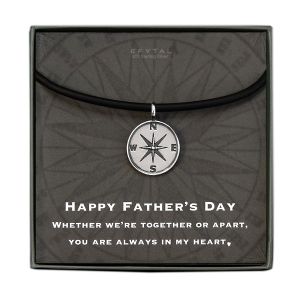 A grey jewelry box containing a grey message card with tonal compass motif. A black silicone cord with a Sterling Silver compass pendant rests on the card. The white text on the card reads “EFYTAL 925 Sterling Silver” at the top and “HAPPY FATHER’S DAY WHETHER WE’RE TOGETHER OR APART, YOU ARE ALWAYS IN MY HEART <3 .”  