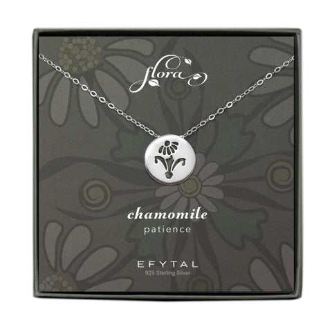 A grey jewelry box contains a grey message card with yellow and green lavender floral motif. A sterling silver necklace with a circular pendant rests on the card. The pendant has a cutout in the shape of a chamomile flower. The text above the necklace reads "flora” and the text below reads “chamomile patience EFYTAL 925 Sterling Silver”