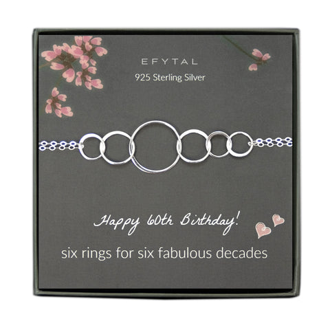 grey jewelry box containing silver bracelet with six thin interlocking circles, placed on a grey card with pink flowers and branches at the top. the text on the card reads "EFYTAL 925 sterling silver" at the top and "Happy 60th Birthday! six rings for six fabulous decades <3 <3" at the bottom.