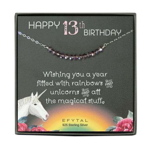 grey jewelry box containing silver necklace with thirteen iridescent purple glass beads, placed on a grey card. The bottom left corner of the card has a unicorn and roses, there is a rainbow in the bottom center, and a rose on the bottom right corner. The card reads "Happy 13th birthday" in pink and white font at the top, and "Wishing you a year filled with rainbows and unicorns and all the magical stuff <3" underneath in white. "EFYTAL 925 sterling silver" is in small dark text at the bottom of the card.