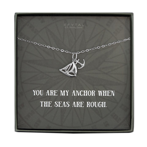 A grey jewelry box containing a grey message card with tonal compass motif. A 925 Sterling Silver necklace with a Sterling Silver pendant with a dangling sailboat and anchor rests on the card. The white text on the card reads “EFYTAL 925 Sterling Silver” at the top and “YOU ARE MY ANCHOR WHEN THE SEAS ARE ROUGH” at the bottom.  