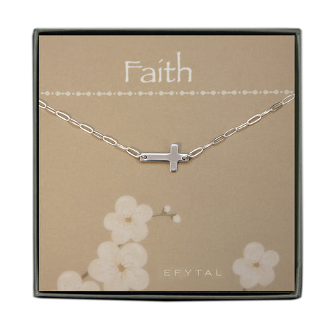 A grey jewelry box containing a tan message card with a white floral motif at the bottom. A silver necklace with a modern chain link and a sideways silver cross pendant rests on the card. The white text on the card reads “Faith” at the top and “EFYTAL” at the bottom.
