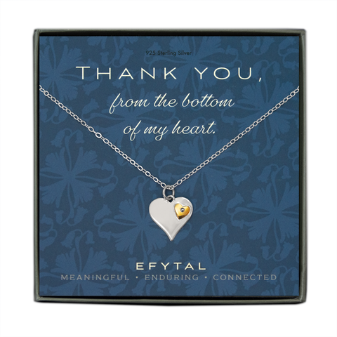 A grey jewelry box golds a blue message card with floral patterned background. A silver necklace rests on the card. The necklace has a heart shaped pendant with a small embedded bronze heart on top. The white text on the card reads “THANK YOU, from the bottom of my heart.” at the top and "EFYTAL MEANINGFUL - ENDURING - CONNECTED” on the bottom.