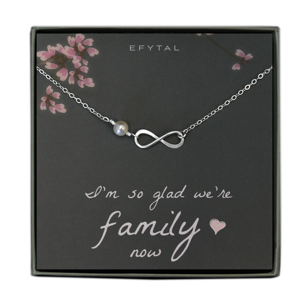 A grey jewelry box contains a grey message card with pink flower and branch motifs at the top. A silver necklace rests on the card - the necklace has an ivory pearl and a silver infinity sign pendant. The white text reads "EFYTAL" at the top and "I'm so glad we're family <3 now" at the bottom.