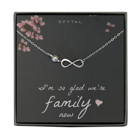 A grey jewelry box contains a grey message card with pink flower and branch motifs at the top. A silver necklace rests on the card - the necklace has an ivory pearl and a silver infinity sign pendant. The white text reads "EFYTAL" at the top and "I'm so glad we're family <3 now" at the bottom.