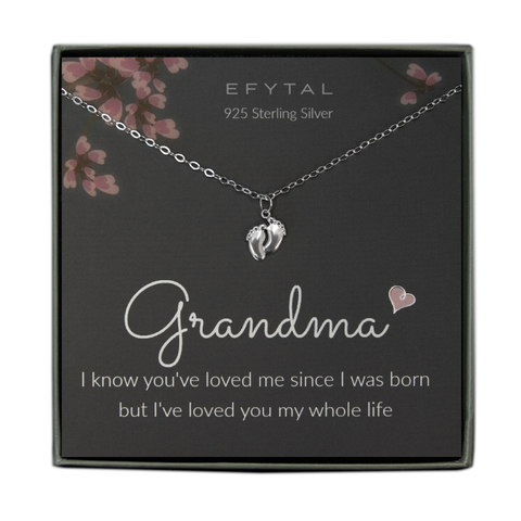A grey jewelry box contains a grey message card with pink flower and branch motif at the top. A sterling silver necklace with a pendant of two tiny feet rests on the card. The white text at the top reads "EFYTAL 925 Sterling Silver" and "Grandma <3 I know you've loved me since I was born but I've loved you my whole life" at the bottom.