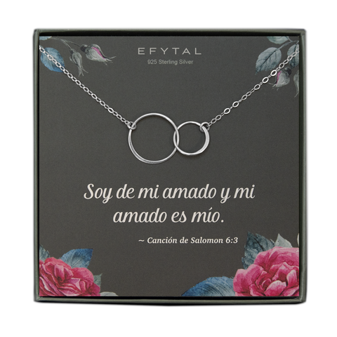 A grey jewelry box containing a grey message card with pink rose motif in the bottom two corners, and rosebuds with leaves in the top two. A sterling silver necklace with a pendant of two interlocking circles rests on the card. The white text on the card reads "EFYTAL" at the top and “Soy de mi amado y mi amado es mio. ~ Canción de Salomon 6:3” at the bottom.