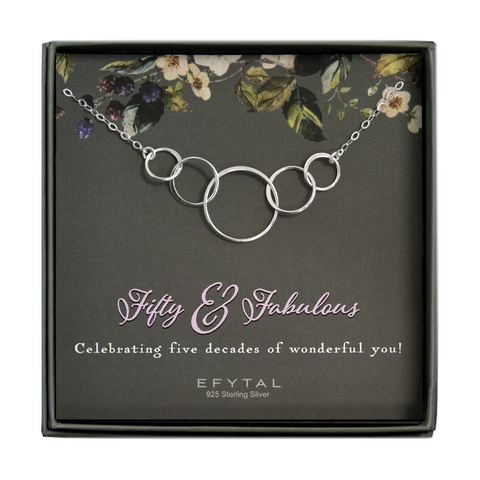 grey jewelry box containing silver necklace with five thin interlocking circles, placed on a grey card with cream and purple flowers and green leaves at the top. the text at the bottom of the card reads "Fifty & Fabulous" in large looping lilac font and "Celebrating fivedecades of wonderful you!" in smaller cream font underneath. At the very bottom: "EFYTAL 925 Sterling Silver" is in small cream font.