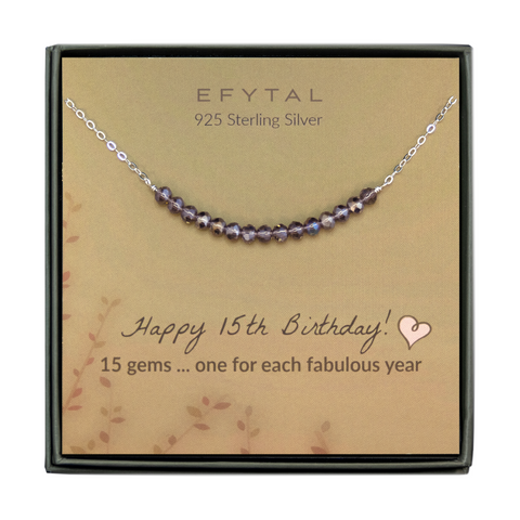  grey jewelry box containing silver necklace with fifteen iridescent purple glass beads, placed on a faded yellow card with a branch motif in the lower left corner. the text on the card reads "EFYTAL 925 sterling silver" in brown at the top and "Happy 15th Birthday! 15 gems...one for each fabulous year" at the bottom.
