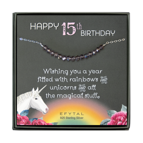 grey jewelry box containing silver necklace with fifteen iridescent purple glass beads, placed on a grey card. The bottom left corner of the card has a unicorn and roses, there is a rainbow in the bottom center, and a rose on the bottom right corner. The card reads "Happy 15th birthday" in pink and white font at the top, and "Wishing you a year filled with rainbows and unicorns and all the magical stuff <3" underneath in white. "EFYTAL 925 sterling silver" is in small dark text at the bottom of the card. 