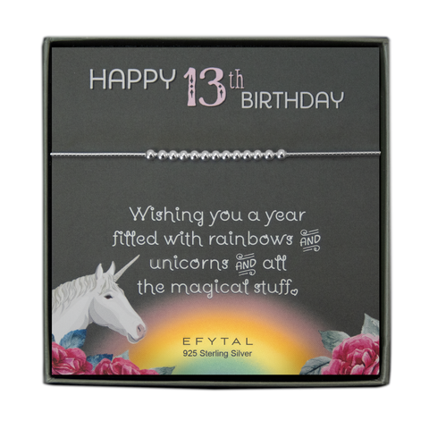 grey jewelry box containing silver bracelet with thirteen round silver beads, placed on a grey card. The bottom left corner of the card has a unicorn and roses, there is a rainbow in the bottom center, and a rose on the bottom right corner. The card reads "Happy 13th birthday" in pink and white font at the top, and "Wishing you a year filled with rainbows and unicorns and all the magical stuff <3" underneath in white. "EFYTAL 925 sterling silver" is in small dark text at the bottom of the card. 