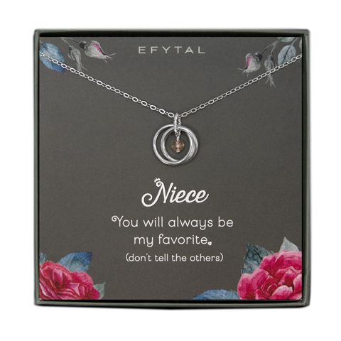 A grey jewelry box containing a grey message card with pink rose motif in the bottom two corners, and rosebuds with leaves in the top two. A sterling silver necklace with two connected silver rings and a dangling champagne stone in the center rests on the card. The white text on the card reads "EFYTAL" at the top and “Niece You will always be my favorite <3 (don’t tell the others)” at the bottom.